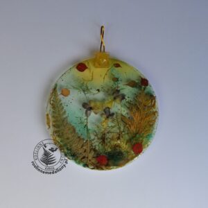 Ręcznie wykonany, okrągły medalion z żywicy, w którym zatopiono różnobarwne suszone rośliny, umieszczony na kartce papieru.