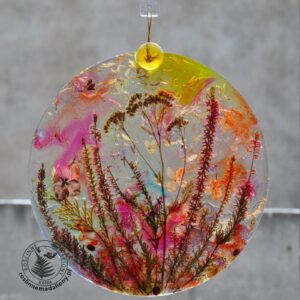 Ręcznie wykonany, okrągły medalion z żywicy, w którym zatopiono różnobarwne suszone rośliny, w tym liście paproci i bratki, zawieszony na szybie okna.