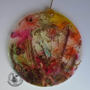 Ręcznie wykonany, okrągły medalion z żywicy, w którym zatopiono różnobarwne suszone rośliny, umieszczony na kartce papieru.