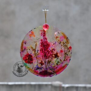 Ręcznie wykonany, okrągły medalion z żywicy, w którym zatopiono różnobarwne suszone rośliny, w tym liście paproci i bratki, zawieszony na szybie okna.