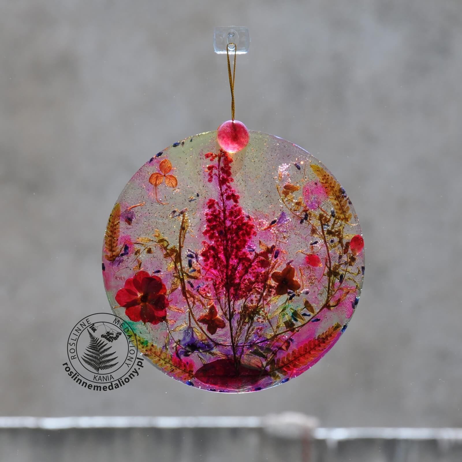 Ręcznie wykonany, okrągły medalion z żywicy, w którym zatopiono różnobarwne suszone rośliny, w tym liście paproci i bratki, zawieszony na szybie okna.
