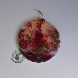 Ręcznie wykonany, okrągły medalion z żywicy, w którym zatopiono różnobarwne suszone rośliny, umieszczony na kartce papieru.