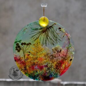 Ręcznie wykonany, okrągły medalion z żywicy, w którym zatopiono różnobarwne suszone rośliny, w tym liście paproci i bratki, zawieszony na szybie okna.
