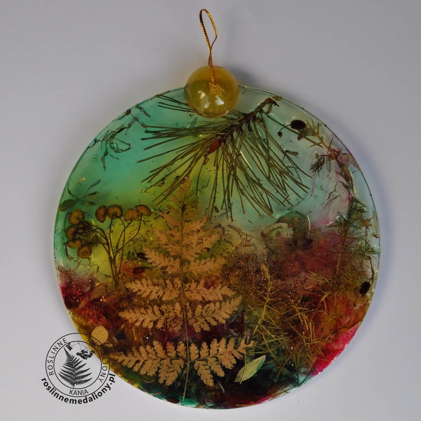 Ręcznie wykonany, okrągły medalion z żywicy, w którym zatopiono różnobarwne suszone rośliny, umieszczony na kartce papieru.