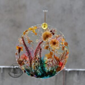 Ręcznie wykonany, okrągły medalion z żywicy, w którym zatopiono różnobarwne suszone rośliny, w tym liście paproci i bratki, zawieszony na szybie okna.