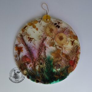 Ręcznie wykonany, okrągły medalion z żywicy, w którym zatopiono różnobarwne suszone rośliny, umieszczony na kartce papieru.