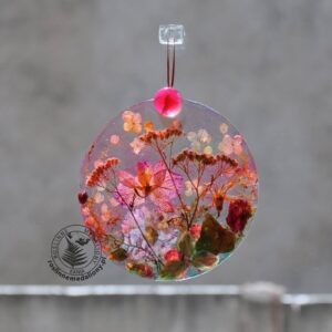 Ręcznie wykonany, okrągły medalion z żywicy, w którym zatopiono różnobarwne suszone rośliny, w tym liście paproci i bratki, zawieszony na szybie okna.