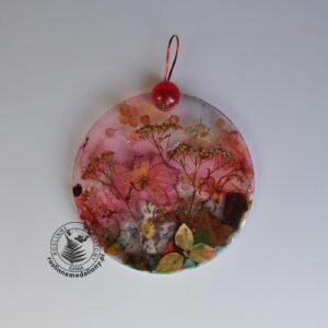 Ręcznie wykonany, okrągły medalion z żywicy, w którym zatopiono różnobarwne suszone rośliny, umieszczony na kartce papieru.