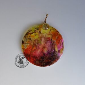 Ręcznie wykonany, okrągły medalion z żywicy, w którym zatopiono różnobarwne suszone rośliny, umieszczony na kartce papieru.