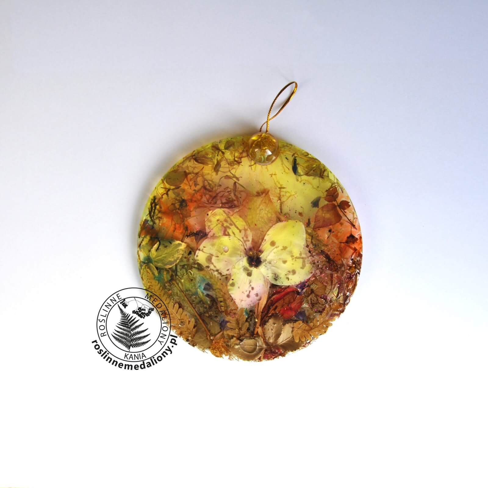 Ręcznie wykonany, okrągły medalion z żywicy, w którym zatopiono różnobarwne suszone rośliny, umieszczony na kartce papieru.