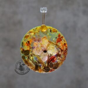 Ręcznie wykonany, okrągły medalion z żywicy, w którym zatopiono różnobarwne suszone rośliny, w tym liście paproci i bratki, zawieszony na szybie okna.