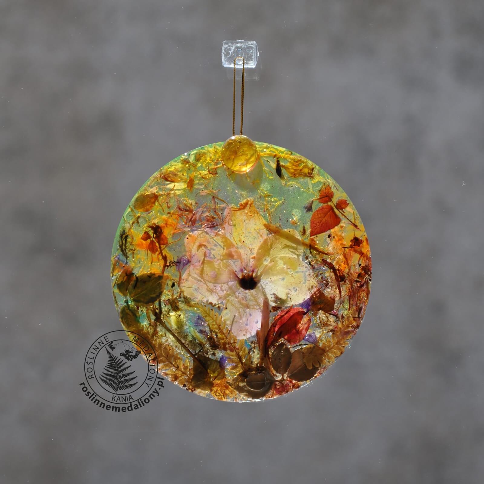 Ręcznie wykonany, okrągły medalion z żywicy, w którym zatopiono różnobarwne suszone rośliny, w tym liście paproci i bratki, zawieszony na szybie okna.