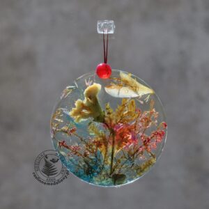 Ręcznie wykonany, okrągły medalion z żywicy, w którym zatopiono różnobarwne suszone rośliny, w tym liście paproci i bratki, zawieszony na szybie okna.