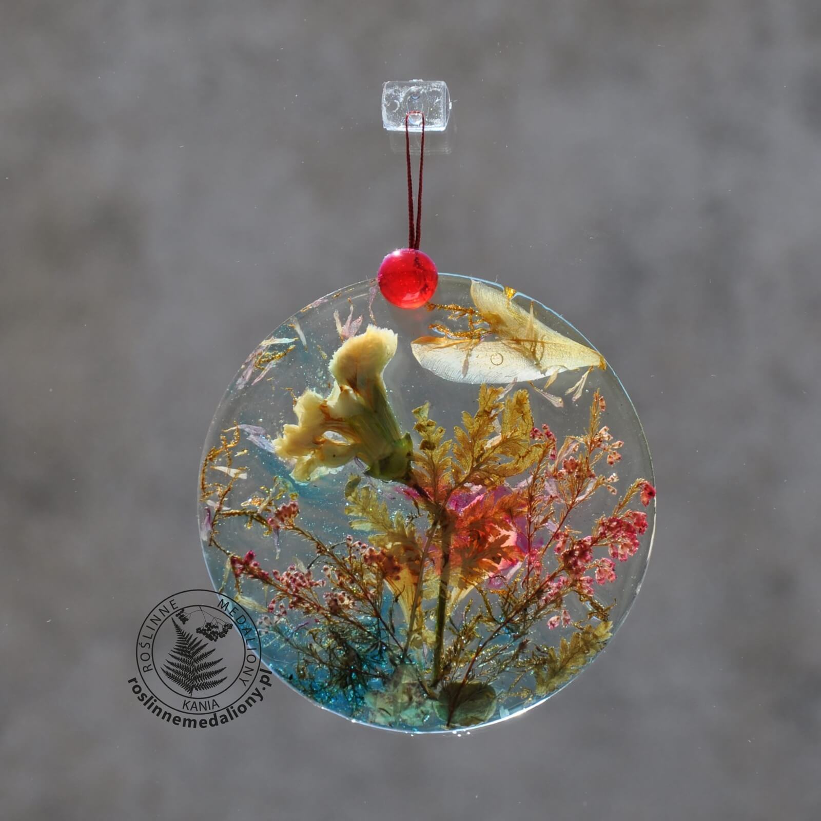 Ręcznie wykonany, okrągły medalion z żywicy, w którym zatopiono różnobarwne suszone rośliny, w tym liście paproci i bratki, zawieszony na szybie okna.