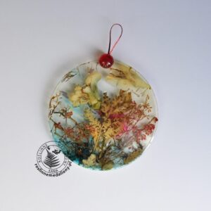 Ręcznie wykonany, okrągły medalion z żywicy, w którym zatopiono różnobarwne suszone rośliny, umieszczony na kartce papieru.