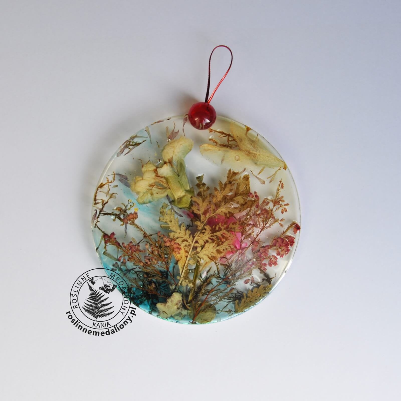 Ręcznie wykonany, okrągły medalion z żywicy, w którym zatopiono różnobarwne suszone rośliny, umieszczony na kartce papieru.