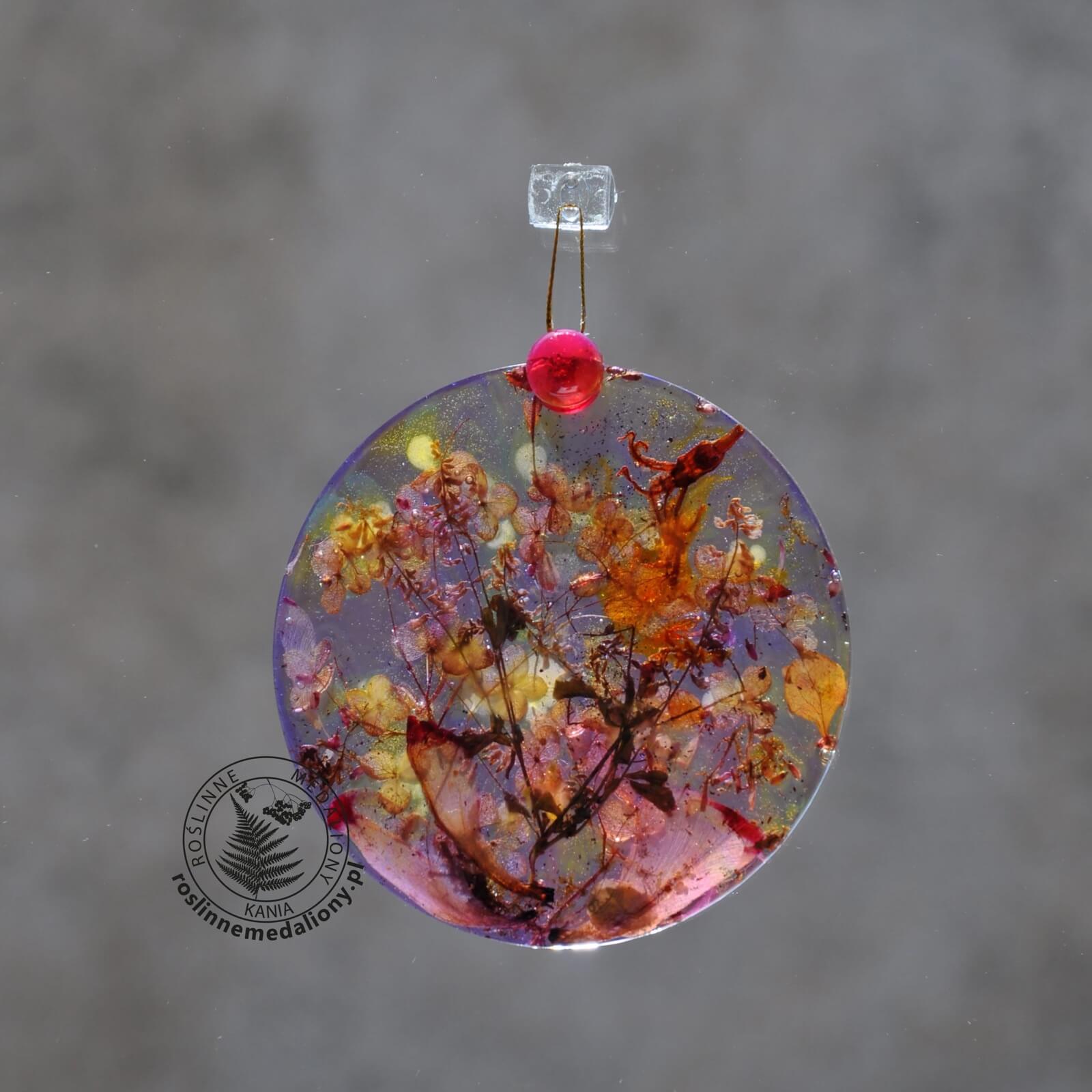 Ręcznie wykonany, okrągły medalion z żywicy, w którym zatopiono różnobarwne suszone rośliny, w tym liście paproci i bratki, zawieszony na szybie okna.