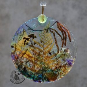 Ręcznie wykonany, okrągły medalion z żywicy, w którym zatopiono różnobarwne suszone rośliny, w tym liście paproci i bratki, zawieszony na szybie okna.