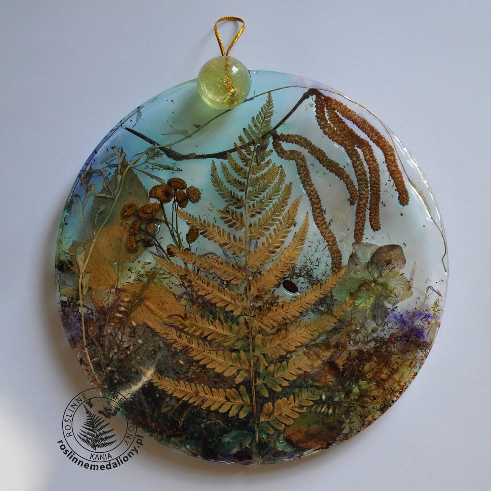 Ręcznie wykonany, okrągły medalion z żywicy, w którym zatopiono różnobarwne suszone rośliny, umieszczony na kartce papieru.