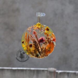 Ręcznie wykonany, okrągły medalion z żywicy, w którym zatopiono różnobarwne suszone rośliny, zawieszony na szybie okna.