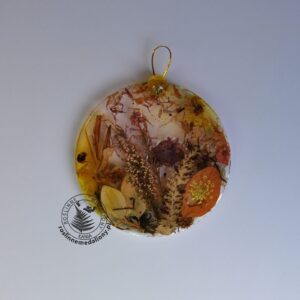 Ręcznie wykonany, okrągły medalion z żywicy, w którym zatopiono różnobarwne suszone rośliny, umieszczony na kartce papieru.