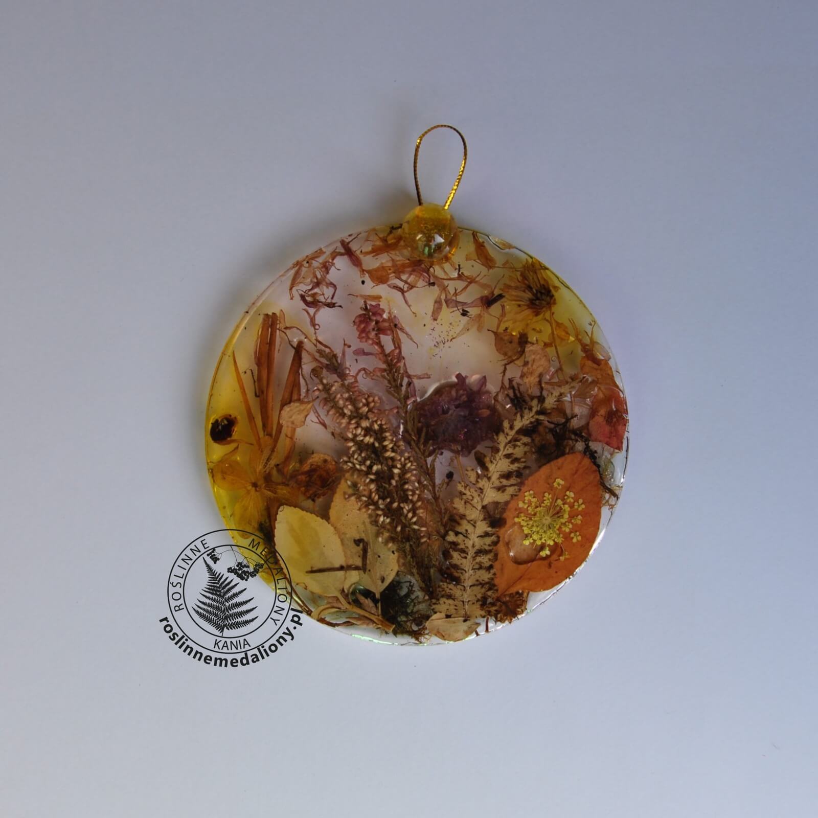 Ręcznie wykonany, okrągły medalion z żywicy, w którym zatopiono różnobarwne suszone rośliny, umieszczony na kartce papieru.