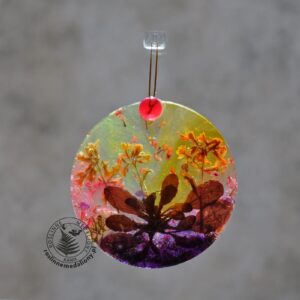 Ręcznie wykonany, okrągły medalion z żywicy, w którym zatopiono różnobarwne suszone rośliny, w tym liście paproci i bratki, zawieszony na szybie okna.