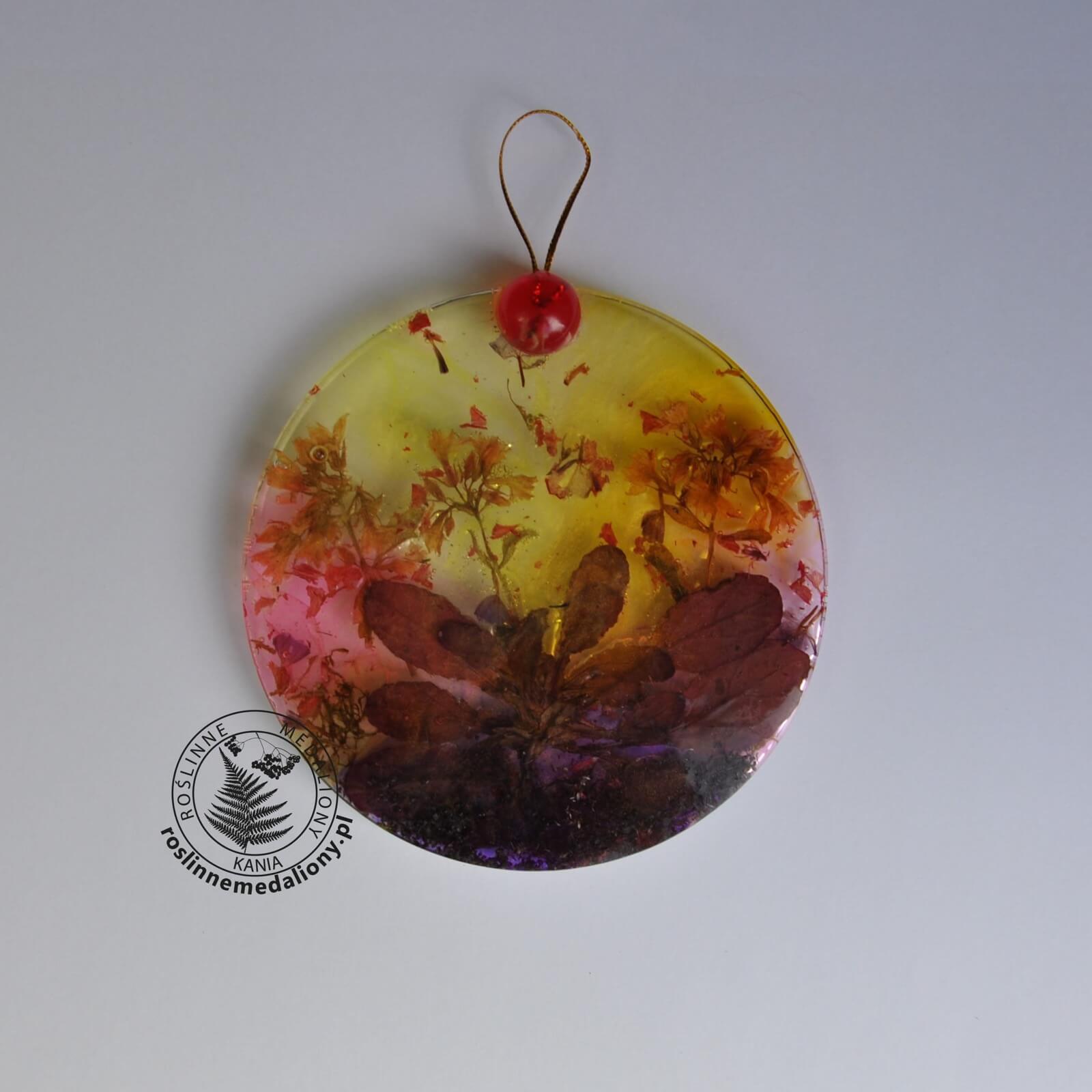 Ręcznie wykonany, okrągły medalion z żywicy, w którym zatopiono różnobarwne suszone rośliny, umieszczony na kartce papieru.