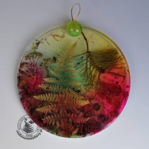 Ręcznie wykonany, okrągły medalion z żywicy, w którym zatopiono różnobarwne suszone rośliny, umieszczony na kartce papieru.