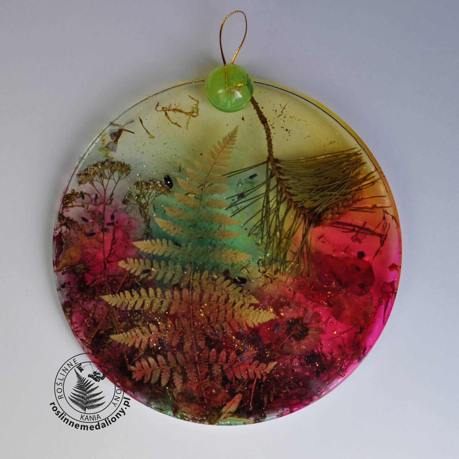 Ręcznie wykonany, okrągły medalion z żywicy, w którym zatopiono różnobarwne suszone rośliny, umieszczony na kartce papieru.