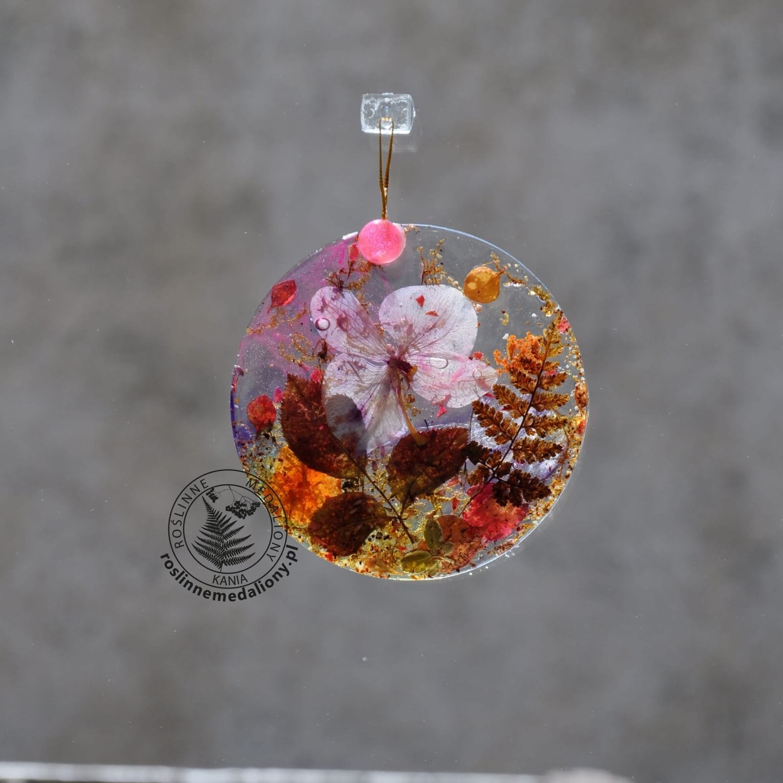 Ręcznie wykonany, okrągły medalion z żywicy, w którym zatopiono różnobarwne suszone rośliny, w tym liście paproci i bratki, zawieszony na szybie okna.