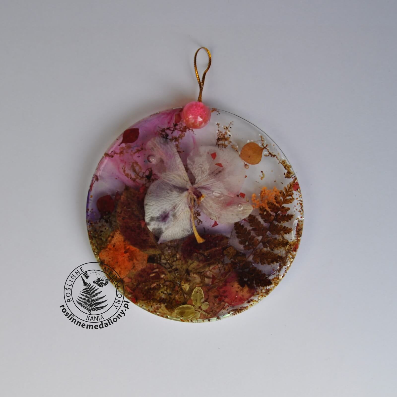 Ręcznie wykonany, okrągły medalion z żywicy, w którym zatopiono różnobarwne suszone rośliny, umieszczony na kartce papieru.