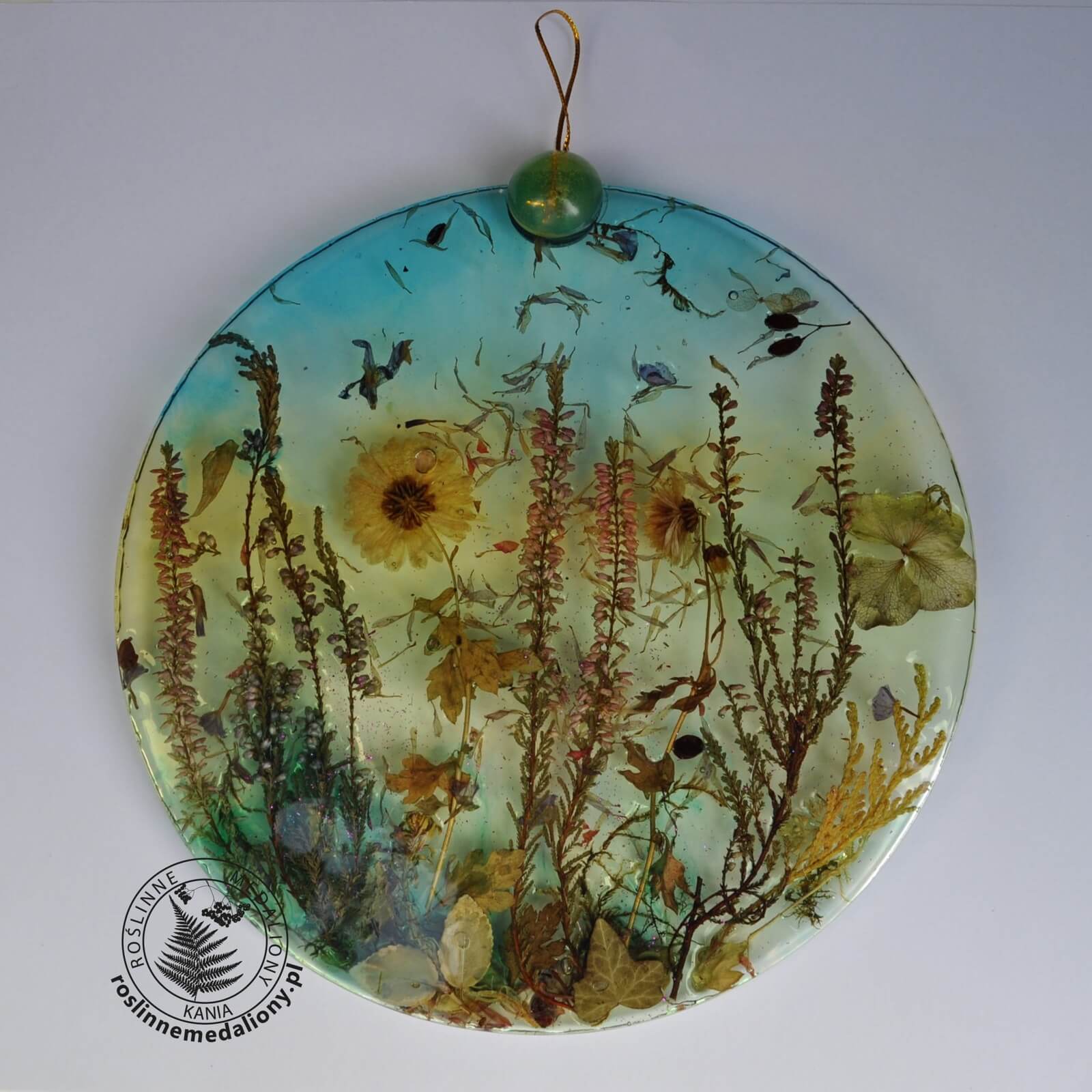 Ręcznie wykonany, okrągły medalion z żywicy, w którym zatopiono różnobarwne suszone rośliny, umieszczony na kartce papieru.