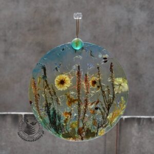 Ręcznie wykonany, okrągły medalion z żywicy, w którym zatopiono różnobarwne suszone rośliny, w tym liście paproci i bratki, zawieszony na szybie okna.