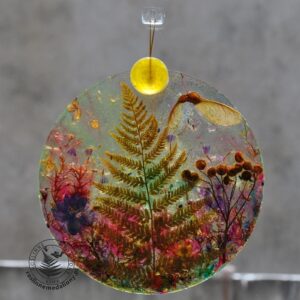 Ręcznie wykonany, okrągły medalion z żywicy, w którym zatopiono różnobarwne suszone rośliny, w tym liście paproci i bratki, zawieszony na szybie okna.