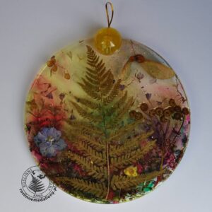 Ręcznie wykonany, okrągły medalion z żywicy, w którym zatopiono różnobarwne suszone rośliny, umieszczony na kartce papieru.