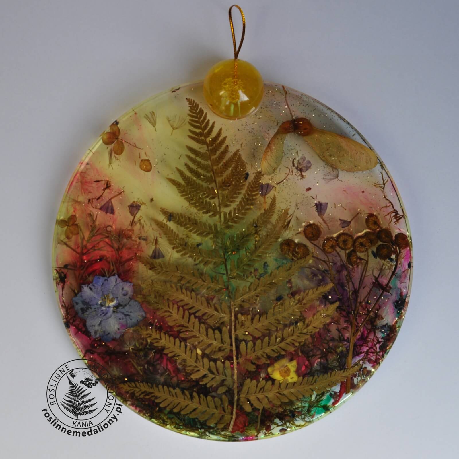 Ręcznie wykonany, okrągły medalion z żywicy, w którym zatopiono różnobarwne suszone rośliny, umieszczony na kartce papieru.