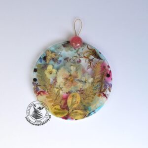 Ręcznie wykonany, okrągły medalion z żywicy, w którym zatopiono różnobarwne suszone rośliny, umieszczony na kartce papieru.