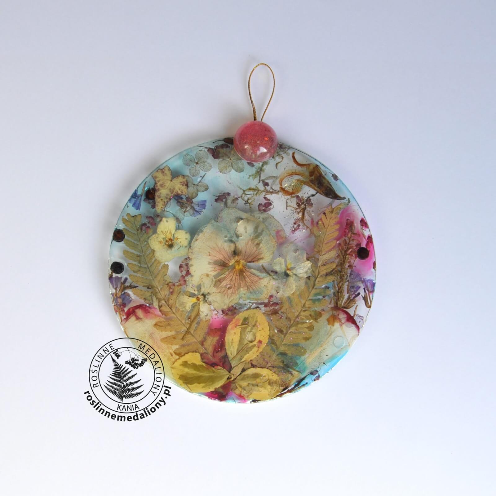 Ręcznie wykonany, okrągły medalion z żywicy, w którym zatopiono różnobarwne suszone rośliny, umieszczony na kartce papieru.