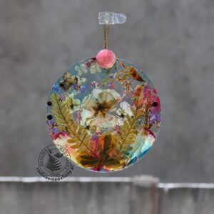 Ręcznie wykonany, okrągły medalion z żywicy, w którym zatopiono różnobarwne suszone rośliny, zawieszony na szybie okna.