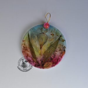 Ręcznie wykonany, okrągły medalion z żywicy, w którym zatopiono różnobarwne suszone rośliny, umieszczony na kartce papieru.