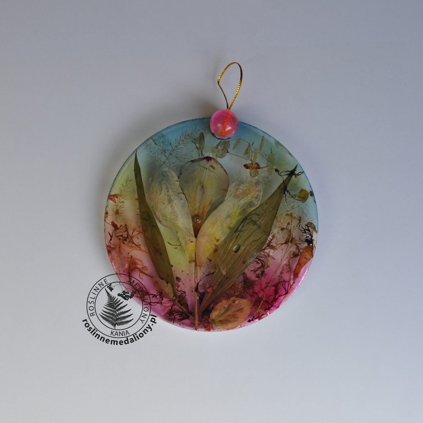 Ręcznie wykonany, okrągły medalion z żywicy, w którym zatopiono różnobarwne suszone rośliny, umieszczony na kartce papieru.
