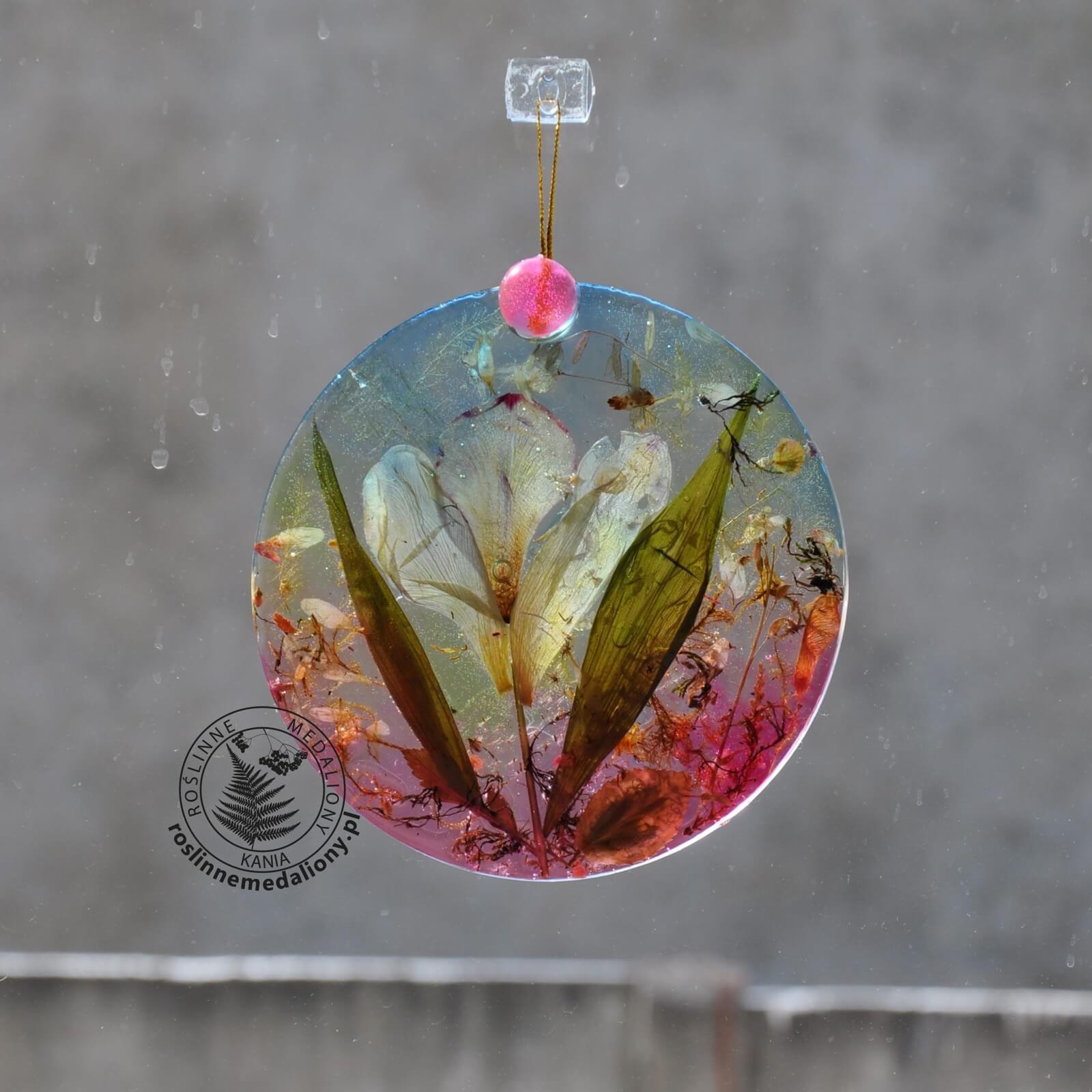 Ręcznie wykonany, okrągły medalion z żywicy, w którym zatopiono różnobarwne suszone rośliny, zawieszony na szybie okna.