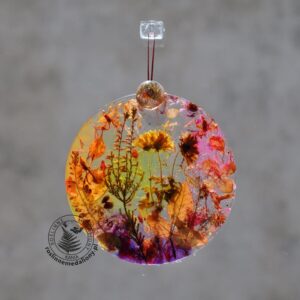 Ręcznie wykonany, okrągły medalion z żywicy, w którym zatopiono różnobarwne suszone rośliny, zawieszony na szybie okna.