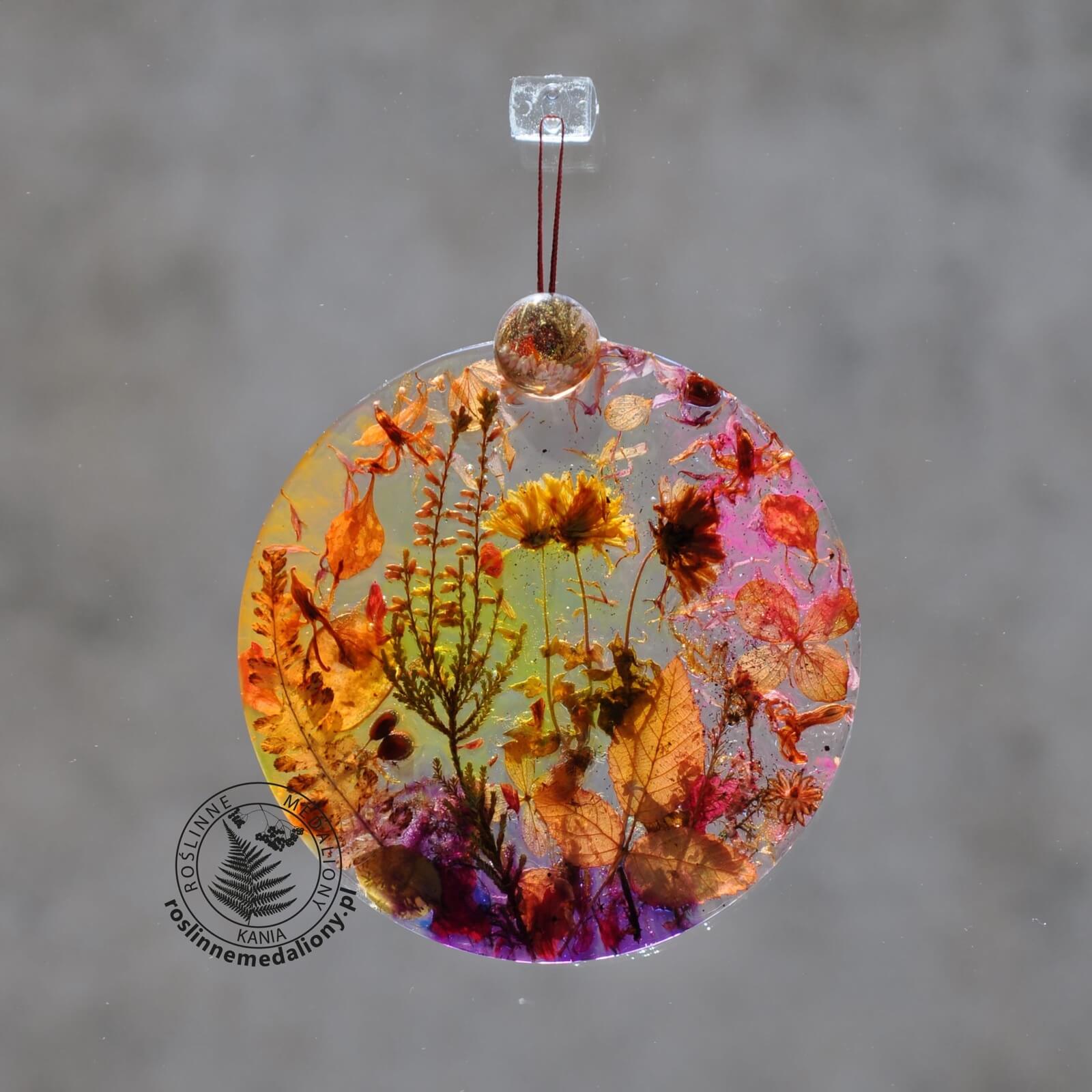 Ręcznie wykonany, okrągły medalion z żywicy, w którym zatopiono różnobarwne suszone rośliny, zawieszony na szybie okna.