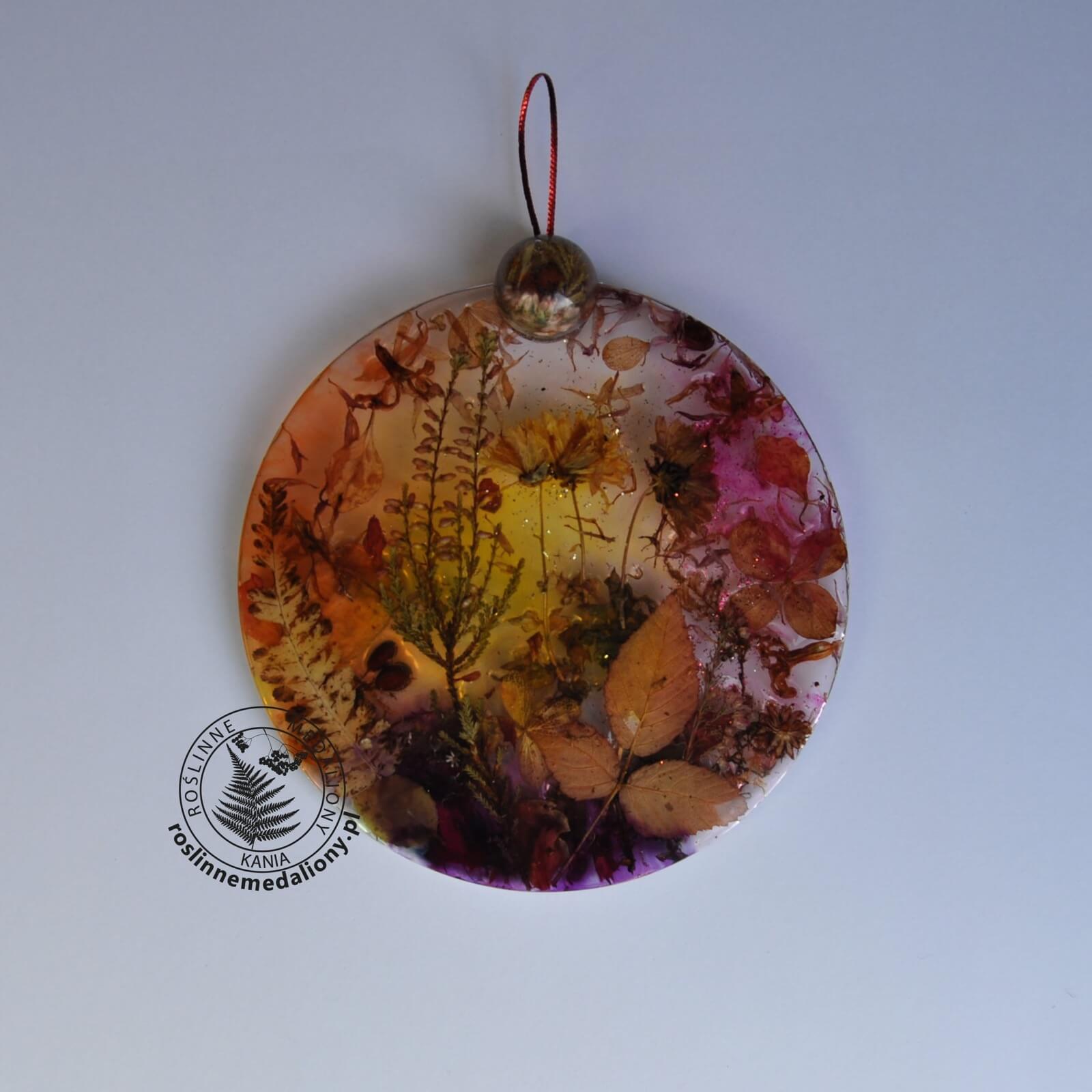 Ręcznie wykonany, okrągły medalion z żywicy, w którym zatopiono różnobarwne suszone rośliny, umieszczony na kartce papieru.