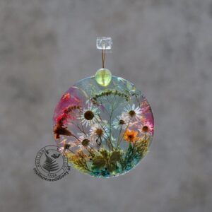 Ręcznie wykonany, okrągły medalion z żywicy, w którym zatopiono różnobarwne suszone rośliny, zawieszony na szybie okna.