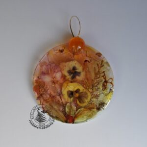 Ręcznie wykonany, okrągły medalion z żywicy, w którym zatopiono różnobarwne suszone rośliny, umieszczony na kartce papieru.