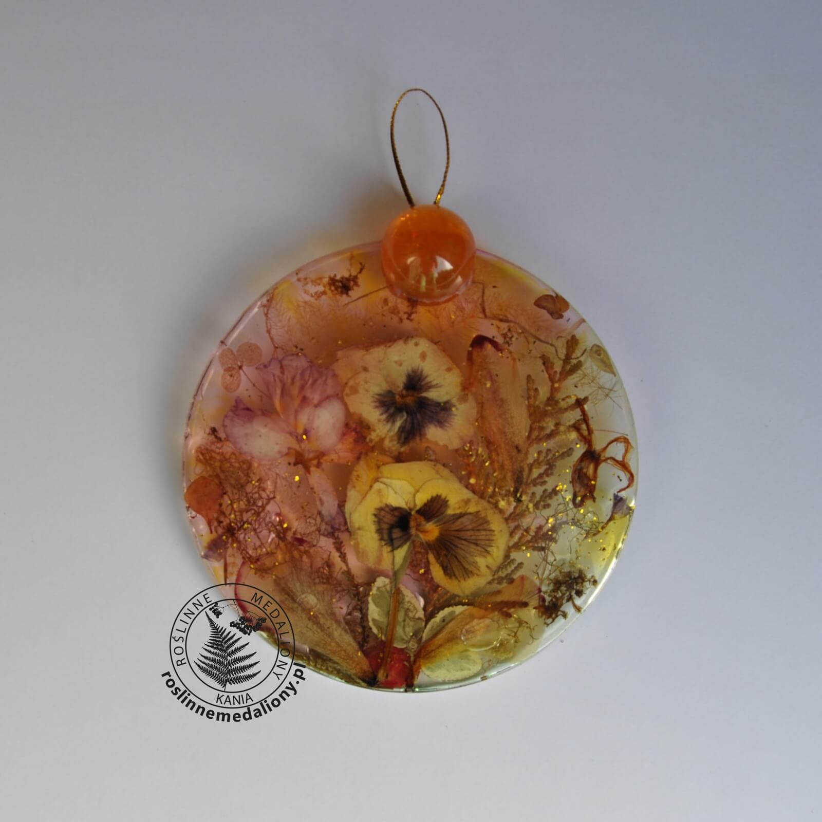 Ręcznie wykonany, okrągły medalion z żywicy, w którym zatopiono różnobarwne suszone rośliny, umieszczony na kartce papieru.