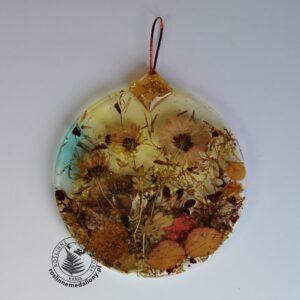 Ręcznie wykonany, okrągły medalion z żywicy, w którym zatopiono różnobarwne suszone rośliny, umieszczony na kartce papieru.